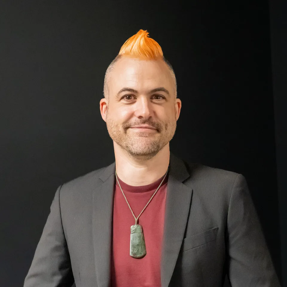 Keri Hēnare smiling, with a bright orange mohawk, a pounamu pendant and a dark blazer