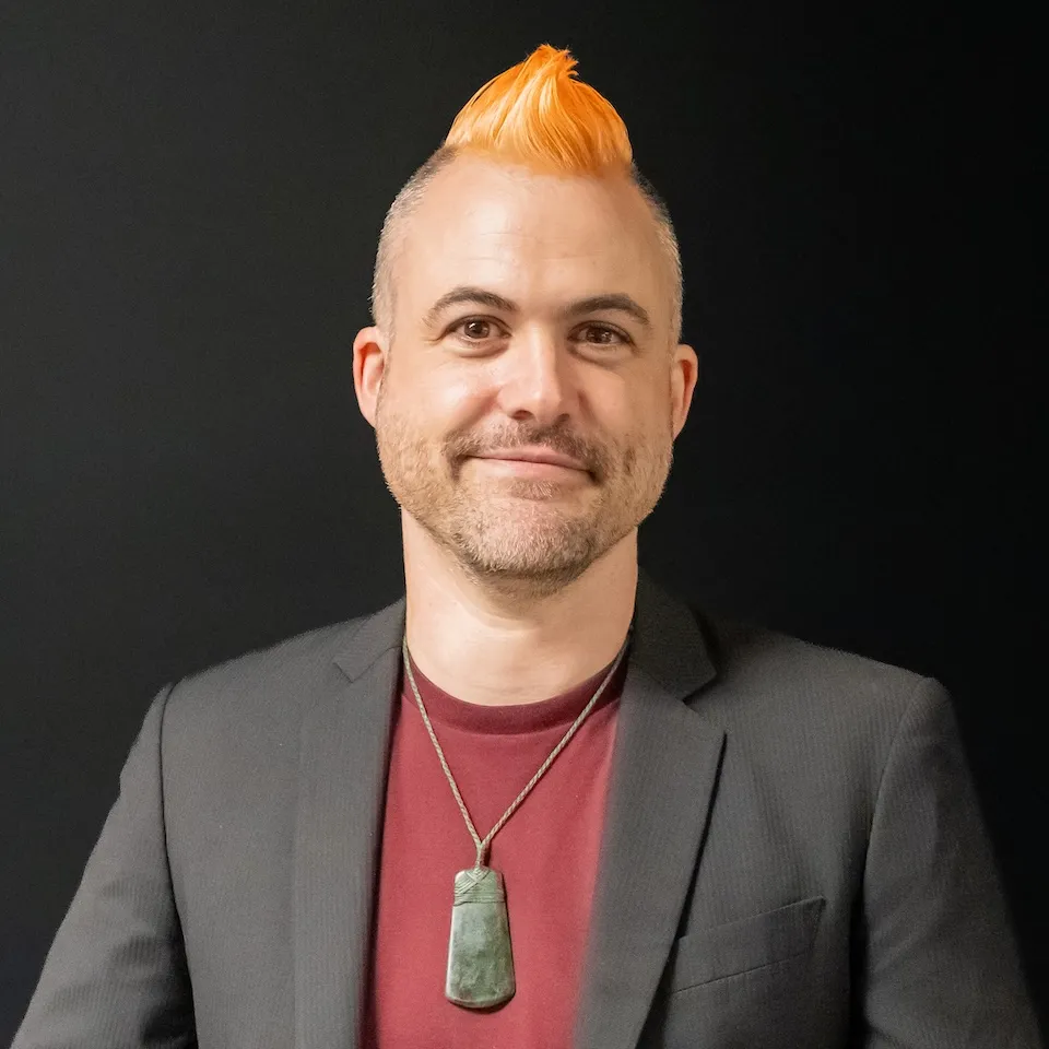 Keri Hēnare smiling, with a bright orange mohawk, a pounamu pendant and a dark blazer